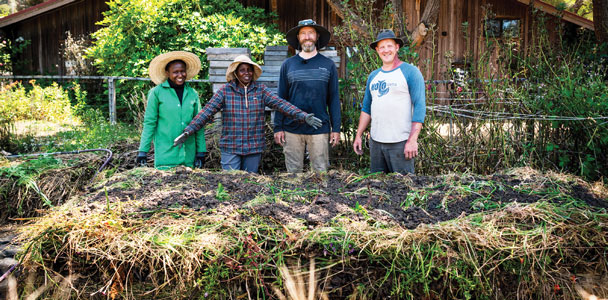 Left to right: Philomena, Teresiah, Matthew and Matt celebrating the construction of a compost pile. Image https://wordofmouthmendo.com/word-of-mouth-stories/2024-fall/the-dance-of-the-elements